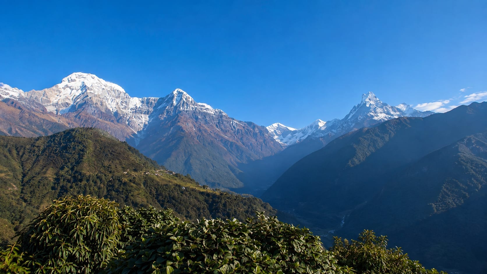 Annapurna South on the left and the fluted spire of Machapuchare on the right, above the forested ridges of the Modi Khola valley, with a scatter of village roofs on the near hillside