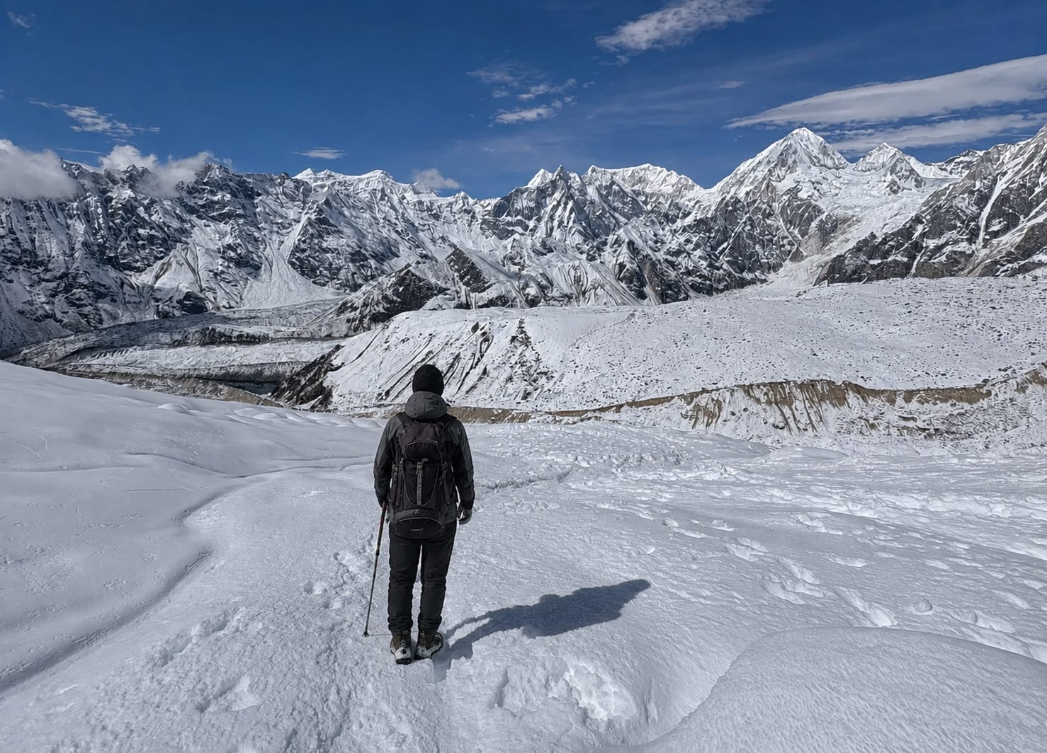 A trekker in a dark jacket walking away across a wide snow-covered glacier, one pole in hand, towards a long wall of snow peaks