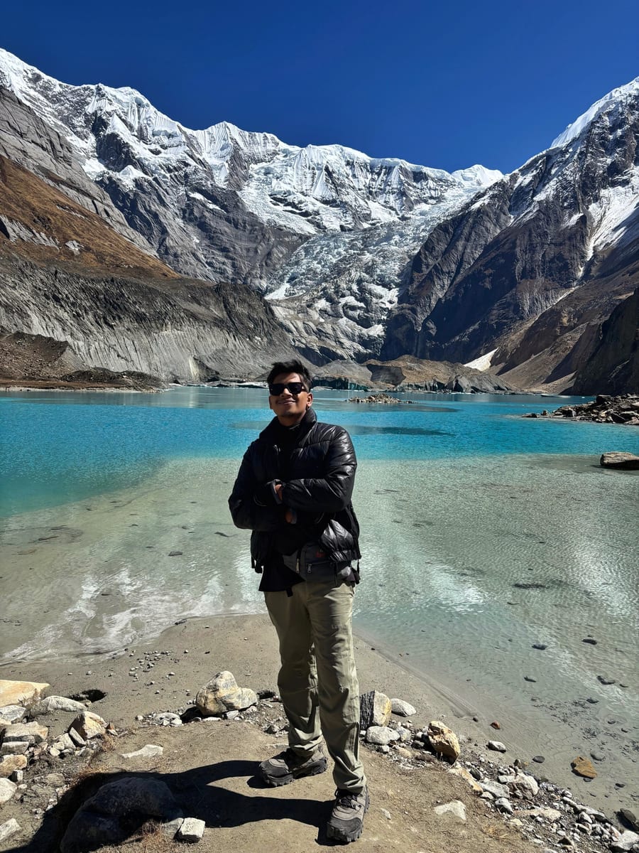 Our founder standing on the gravel shore of the turquoise Panchakunda lake, arms folded, with the glacier and the snow face above the lake behind him