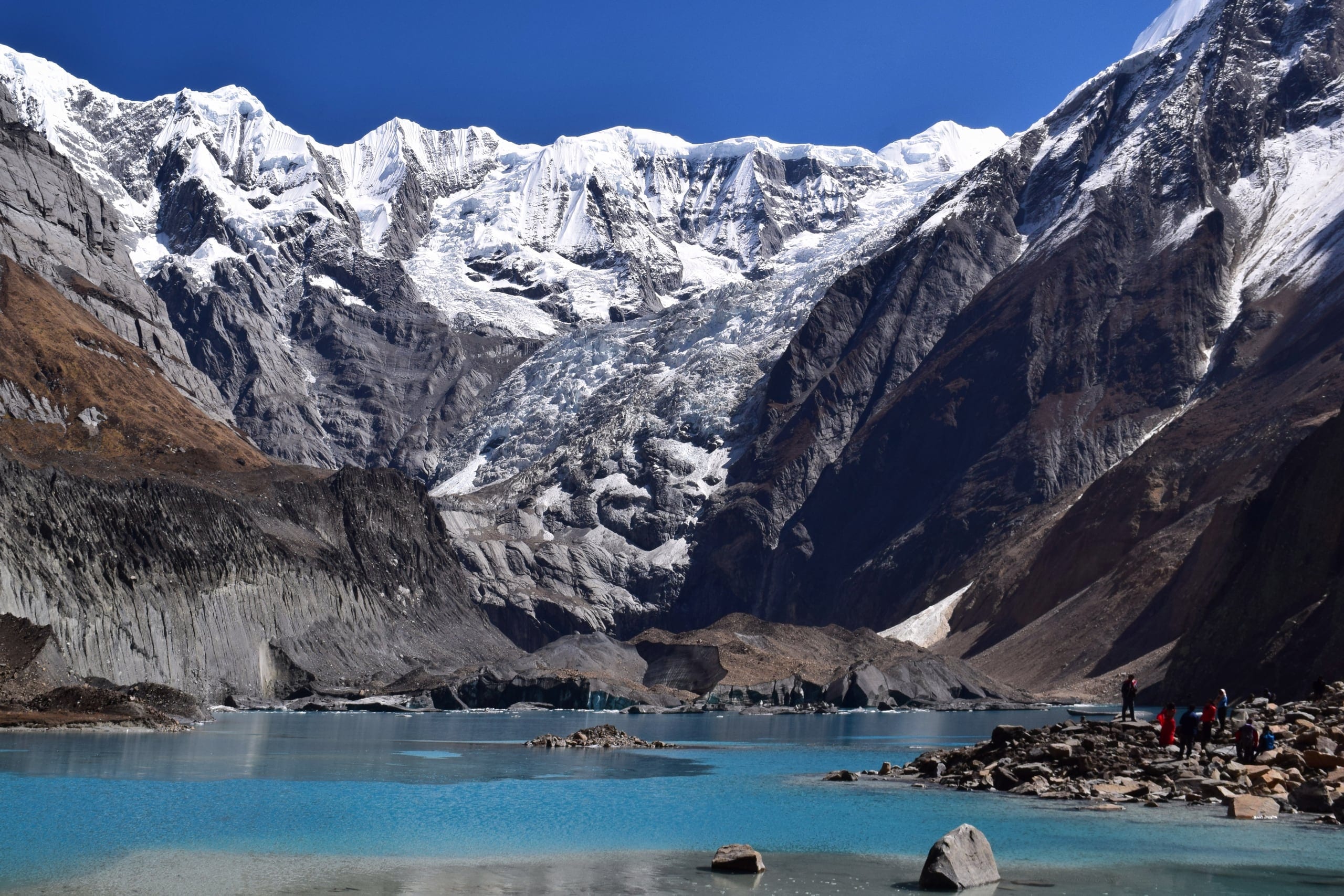 The turquoise Panchakunda lake below the glaciated north face of Annapurna, with trekkers on the rocky shore at the right for scale